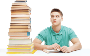 Photo of male student reading with large stack of books next to him - Say the Word ‘Survey’: Part 2 of 2—A Second Look at the Data on Audiology Education Article