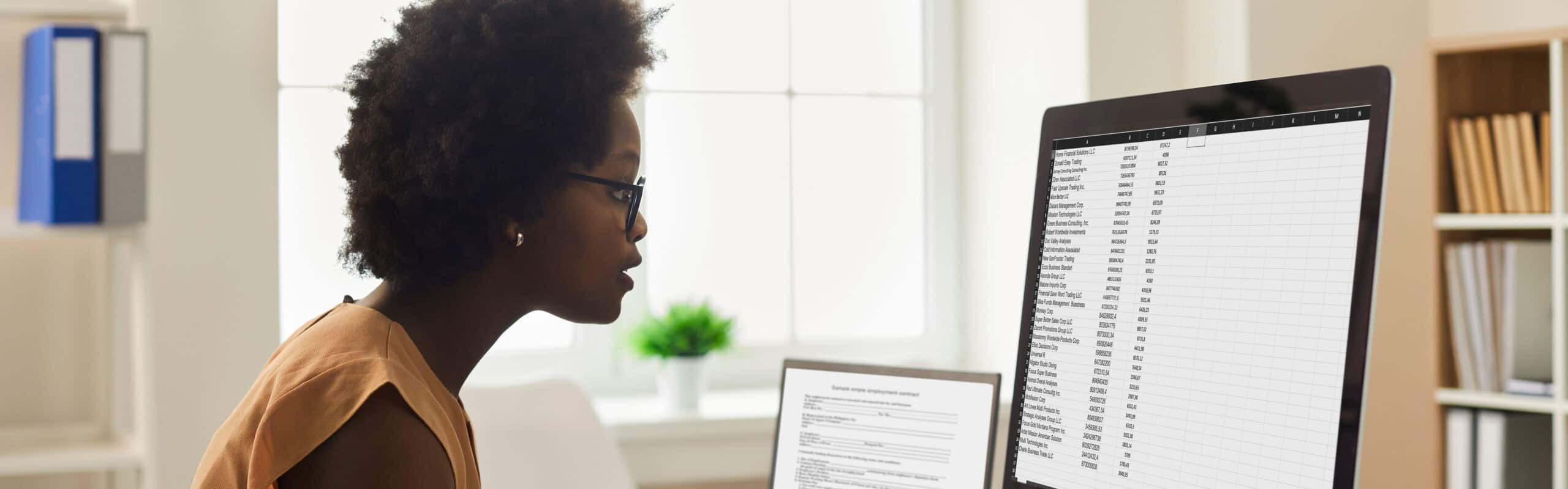 Employee using office computer. African-American woman sitting at desk, looking at desktop PC screen, doing research, studying data reports, working with business documents and online spreadsheets. Shutterstock Image.