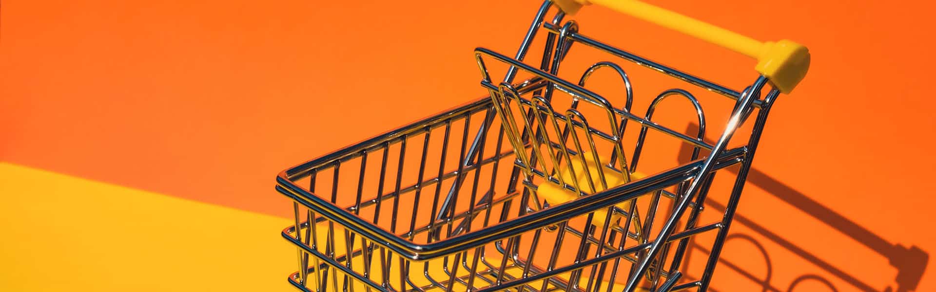 Empty shopping trolley cart on colorful orange yellow background. Copy space for your text. Online shopping, buy mall market shop consumer concept. Small toy supermarket grocery push cart. Food crisis. Shutterstock photo.