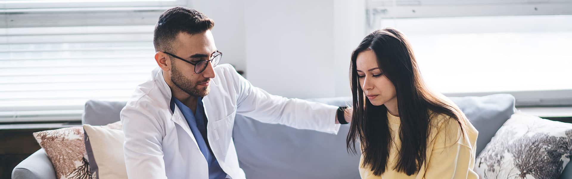 Serious male oncologist in special uniform and glasses carefully touching shoulder of female client after informing about diagnosis while sitting on sofa in office