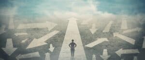 Doubtful person, hands on hips, choosing the way as multiple arrows on the road showing a mess of different directions. Choosing the correct pathway, difficult decision concept, confusion symbol. (StunningArt/Shutterstock.com)