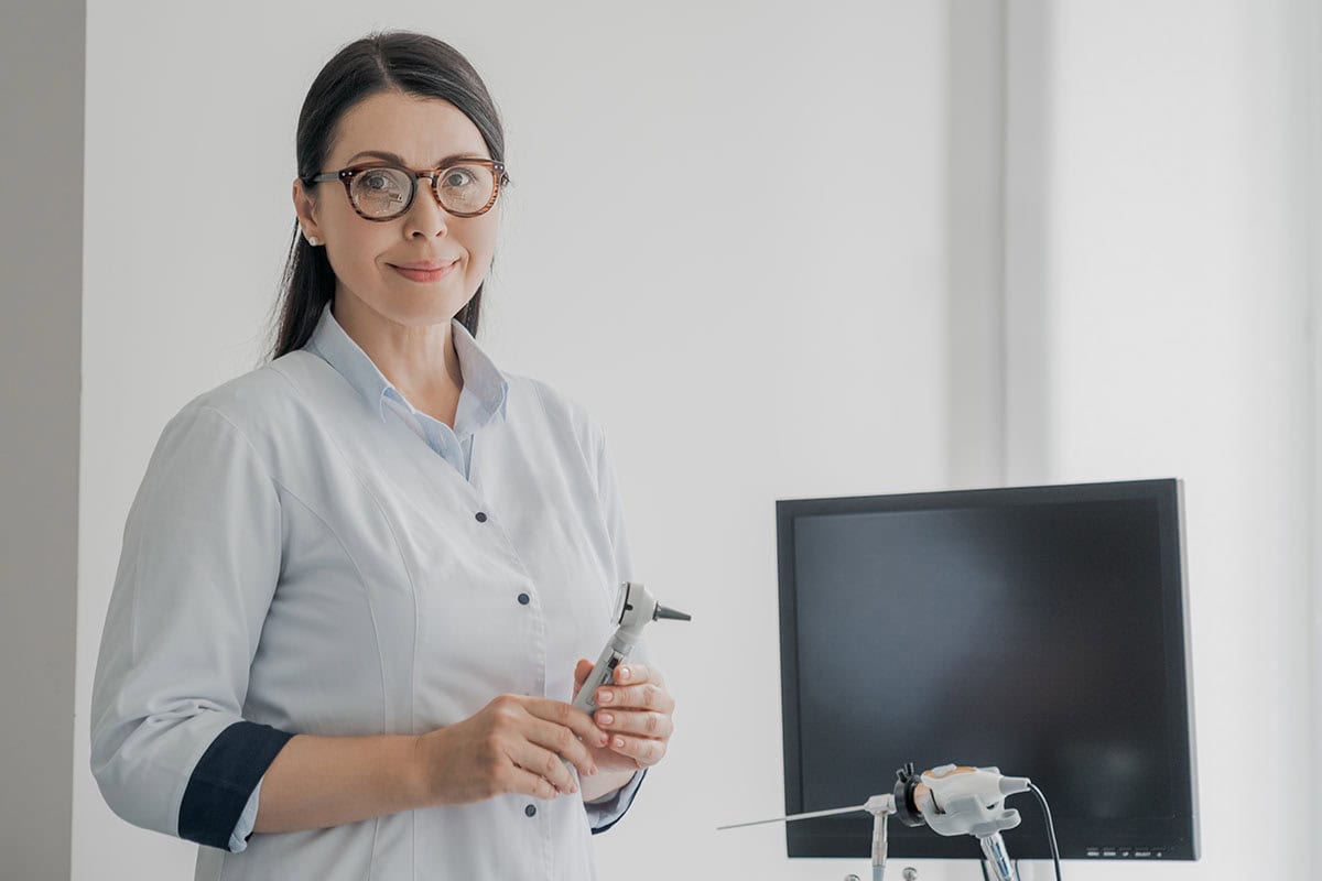 Portrait,Of,Middle-aged,Female,Otolaryngologist,Specialist,With,Otoscope,In,Hands Portrait of middle-aged female otolaryngologist specialist with otoscope in hands looking in camera in modern clinic hospital. Eye nose illnesses treatment.