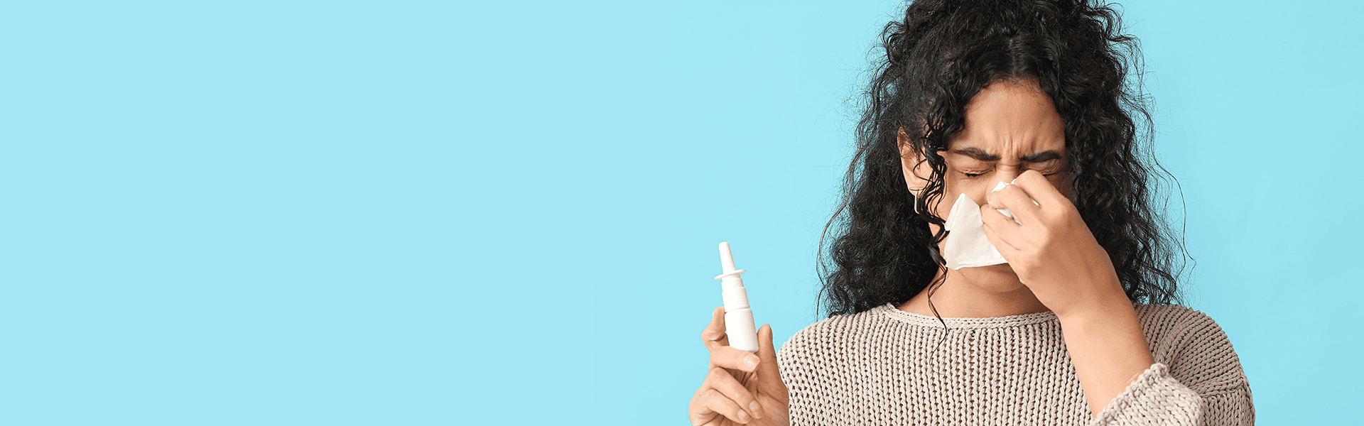Young African-American woman with tissue and nasal spray suffering from allergy on light blue background with space for text