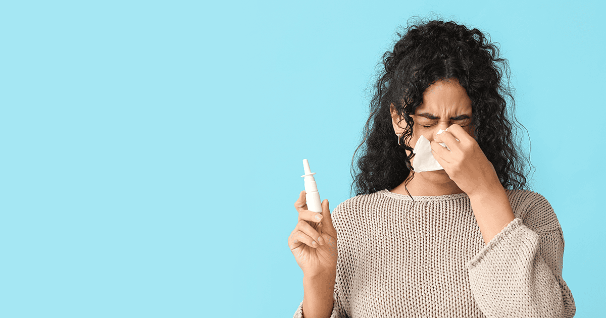 Young African-American woman with tissue and nasal spray suffering from allergy on light blue background with space for text