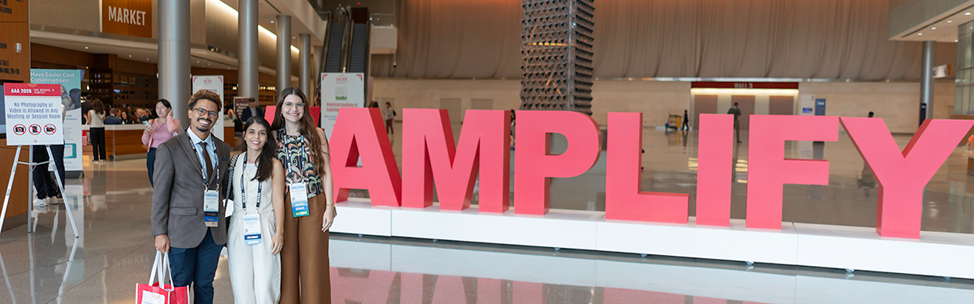 Three attendees posing in a large, modern lobby next to a giant red "AMPLIFY" sign, which is the theme for the AAA 2026 conference.