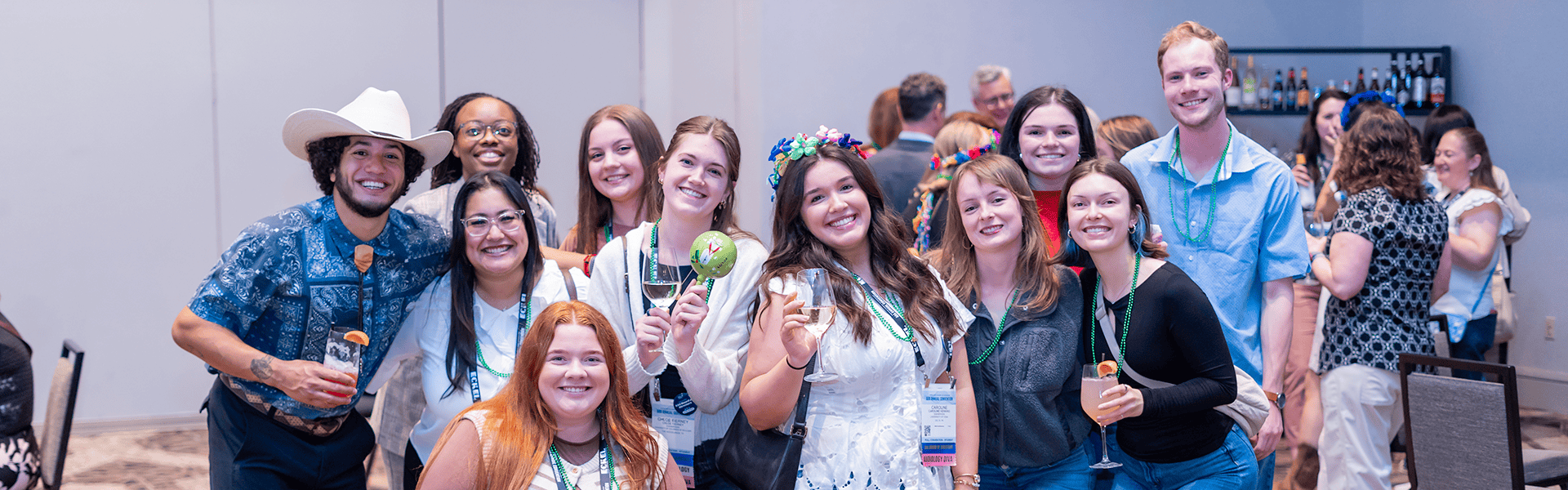 A group of young audiology professionals wearing conference lanyards and festive Mardi Gras beads pose together at the AAA Foundation Fiesta during AAA 2026
