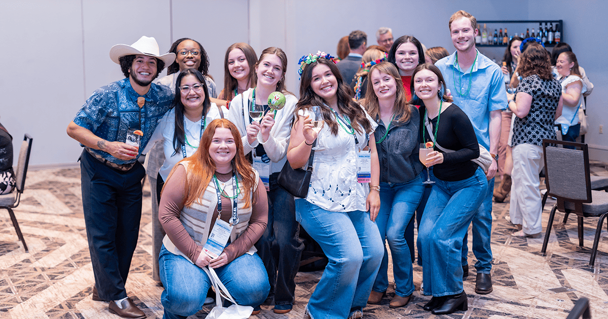 A group of young audiology professionals wearing conference lanyards and festive Mardi Gras beads pose together at the AAA Foundation Fiesta during AAA 2026