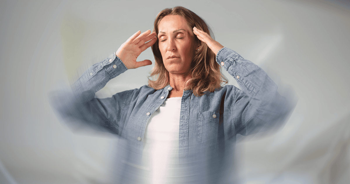 A middle-aged woman with shoulder-length blonde hair stands with her eyes closed and both hands raised to her temples, suggesting dizziness or vestibular disturbance, against a blurred gray background with a swirling motion effect.