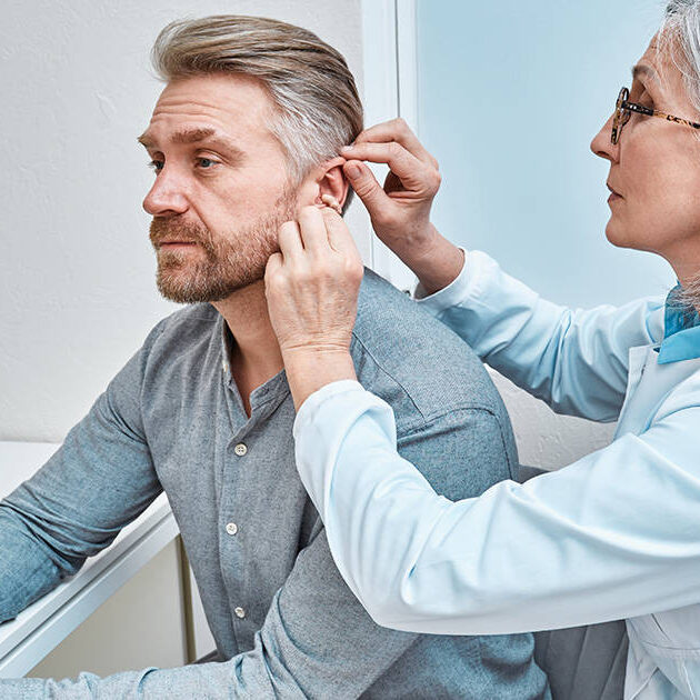 shutterstock_2258166725 - social Experienced female doctor fitting Intra-The-Ear hearing aid into patient's ear while consultation in audiology clinic. ITE and ITC hearing aids